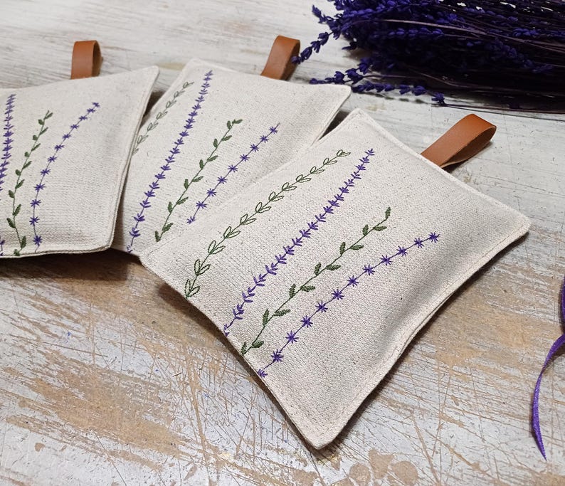 May include: Three beige linen sachets with lavender embroidery. The sachets are filled with dried lavender and are perfect for adding a touch of fragrance to any room.