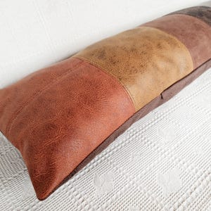 May include: A long rectangular lumbar pillow featuring a patchwork design of faux leather panels in varying shades of brown, including tan, rust orange, and dark chocolate brown, with a visible textured grain pattern across the surface.