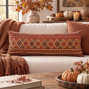 May include: A rectangular lumbar pillow featuring a woven pattern of repeating diamonds and geometric shapes in shades of burnt orange, deep red, and mustard yellow. The fabric has a textured appearance with a consistent, symmetrical design.