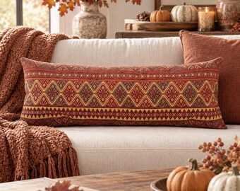 May include: A rectangular lumbar pillow featuring a woven pattern of repeating diamonds and geometric shapes in shades of burnt orange, deep red, and mustard yellow. The fabric has a textured appearance with a consistent, symmetrical design.