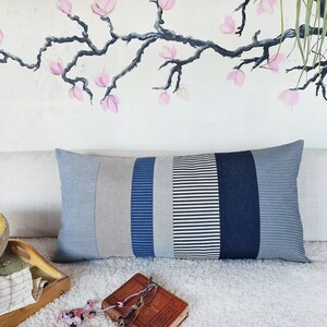 May include: A long rectangular pillow with a patchwork design. The pillow is made of different shades of blue and gray striped fabric. The pillow is sitting on a white fluffy surface.
