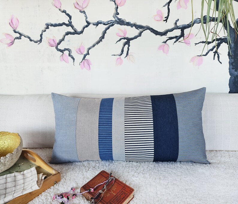 May include: A long rectangular pillow with a patchwork design. The pillow is made of different shades of blue and gray striped fabric. The pillow is sitting on a white fluffy surface.