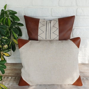May include: A beige pillow with brown leather trim and a geometric pattern on the front. The pillow is sitting on a white brick wall.
