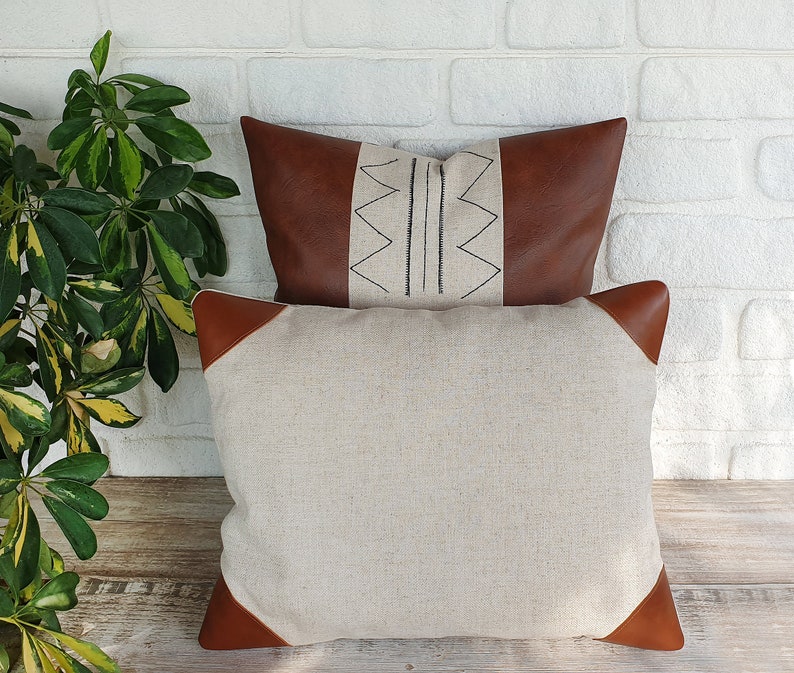 May include: A beige pillow with brown leather trim and a geometric pattern on the front. The pillow is sitting on a white brick wall.