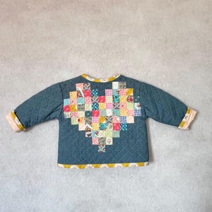 May include: A quilted jacket in a teal blue color with a heart-shaped patchwork design on the back. The patchwork heart is made of various colorful fabric squares. The jacket has a yellow trim and quilted pattern.