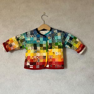 May include: A long-sleeved jacket constructed from a patchwork of small, multicoloured fabric squares in a spectrum of colours. The jacket features a V-neckline, two flower-shaped wooden buttons for closure, and a white lining with colourful dots.