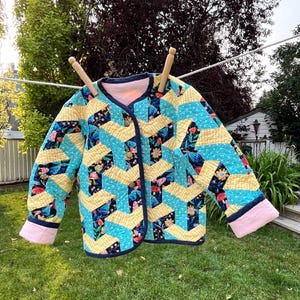 Peut inclure: Une veste matelassée présentant un motif géométrique de tissu vichy jaune et turquoise à pois blancs, entremêlé d'un imprimé floral sombre avec des champignons et des fleurs. La veste a une doublure rose, un liseré bleu foncé et trois boutons.