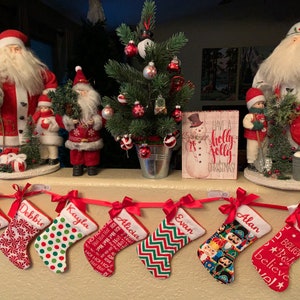May include: A garland of personalized Christmas stockings with red and white ribbon bows. The stockings are decorated with various patterns and designs, including polka dots, stripes, and a nutcracker. The stockings are hung on a fireplace mantel with a small Christmas tree, a snowman figurine, and Santa Claus figurines.