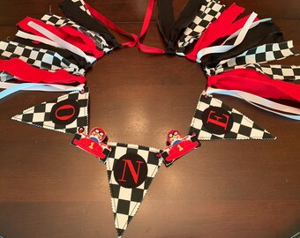May include: A black and white checkered fabric banner with red accents and the word "ONE" spelled out in black circles. The banner is decorated with red and black checkered fabric strips and red ribbon.