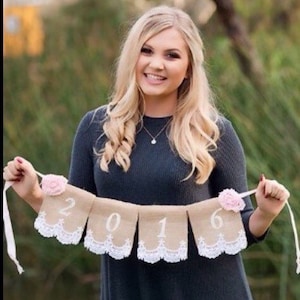 May include: A woman in a gray dress holds a burlap banner with the year 2016 written on it. The banner is decorated with pink flowers and white lace.