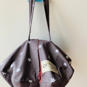 Peut inclure: Un sac fourre-tout en tissu gris avec des étoiles blanches imprimées dessus. Le sac a une fermeture éclair et une étiquette en cuir marron qui dit "Homemade by Rebecca".