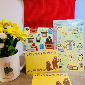 Peut inclure: Deux cartes jaunes avec une illustration d'un ours brun et une bordure de hérissons. Les cartes sont sur une surface en bois avec une tenture murale en tissu rouge, un papier à motifs jaunes et verts, une tasse blanche avec une illustration de chat et une feuille d'autocollants avec un personnage jaune.
