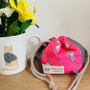 Peut inclure: Un sac à cordon rose avec un imprimé de chat gris et blanc. Le sac a une étiquette blanche qui dit "Handmade By Rebecca".