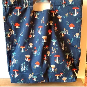 Peut inclure: Un sac fourre-tout bleu marine avec un motif répété de champignons colorés et de feuillage. Les champignons ont des chapeaux rouges, oranges, blancs et beiges. Le sac est de forme rectangulaire avec une grande ouverture et deux poignées.