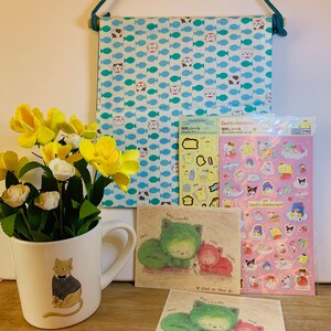 Peut inclure: Un ensemble de quatre articles sur le thème du chat. L'ensemble comprend une suspension murale en tissu avec un motif de poissons bleu et blanc, une tasse en céramique blanche avec une illustration de chat, une feuille d'autocollants avec des personnages Sanrio et deux cartes postales avec des illustrations de chats.