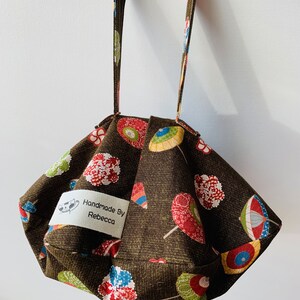 Peut inclure: Un sac en tissu marron avec un motif répété de parasols colorés et de motifs floraux. Le sac porte une étiquette rectangulaire avec l'inscription "Handmade By Rebecca". Le sac a une poignée.
