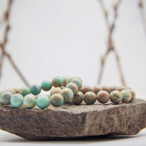 May include: Two turquoise and brown gemstone bracelets stacked on a gray rock. The bracelets are made of round beads.