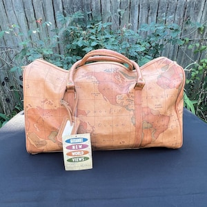 May include: A tan duffel bag featuring a printed world map pattern with two top handles and a rectangular tag attached to the front that reads GRAND NEW WORLD VIEWS in colorful text. The bag has a structured shape with visible stitching details.