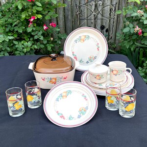 May include: A vintage Corelle dinnerware set featuring a casserole dish, plates, cups, a sugar bowl, and drinking glasses. The set has a white background with a fruit and floral design and a red rim. The casserole dish has a brown lid.