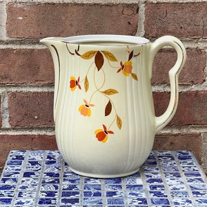 May include: A vintage cream-colored pitcher with a floral design featuring brown leaves and orange flowers. The pitcher has a gold rim and a gold handle.