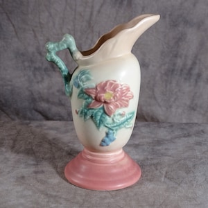 May include: A vintage ceramic pitcher with a floral design. The pitcher is cream colored with a pink base and features a large pink flower with green leaves. The handle is a twisted green branch.