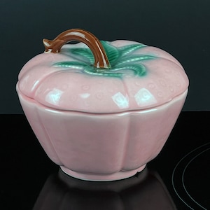 May include: A pink ceramic pumpkin-shaped covered dish with a green leaf and brown stem handle.