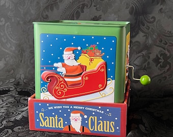 May include: A green metal jack-in-the-box featuring a graphic of Santa Claus in a red sleigh on a blue snowy background. The base is red with the text WE WISH YOU A MERRY CHRISTMAS SANTA CLAUS and a Santa illustration. It has a green crank handle.
