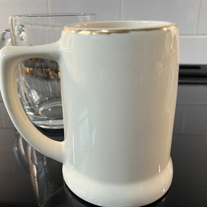 May include: A white ceramic beer stein with a gold rim. The stein has a handle and is sitting on a black surface.