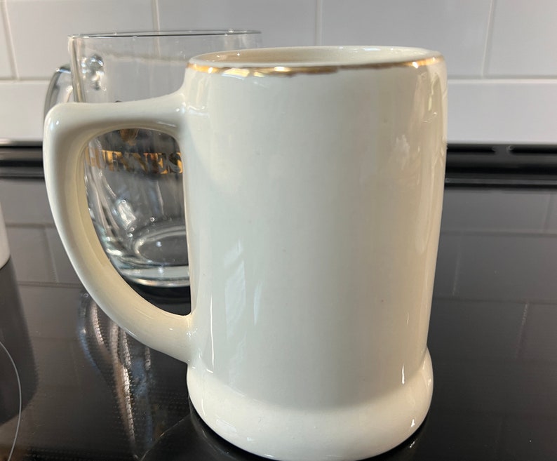 May include: A white ceramic beer stein with a gold rim. The stein has a handle and is sitting on a black surface.