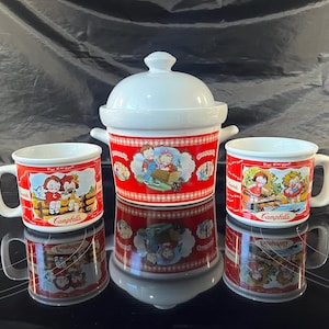 May include: Three white ceramic mugs with red and white designs featuring illustrations of children. The mugs are decorated with the Campbell's Soup logo and the text "Campbells". The mugs are arranged in a triangle shape on a black surface.
