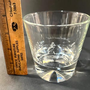 May include: A clear, lowball glass with the Johnnie Walker logo etched on the front. The glass has a thick base and a slightly tapered shape. A wooden ruler with inch markings is next to the glass.