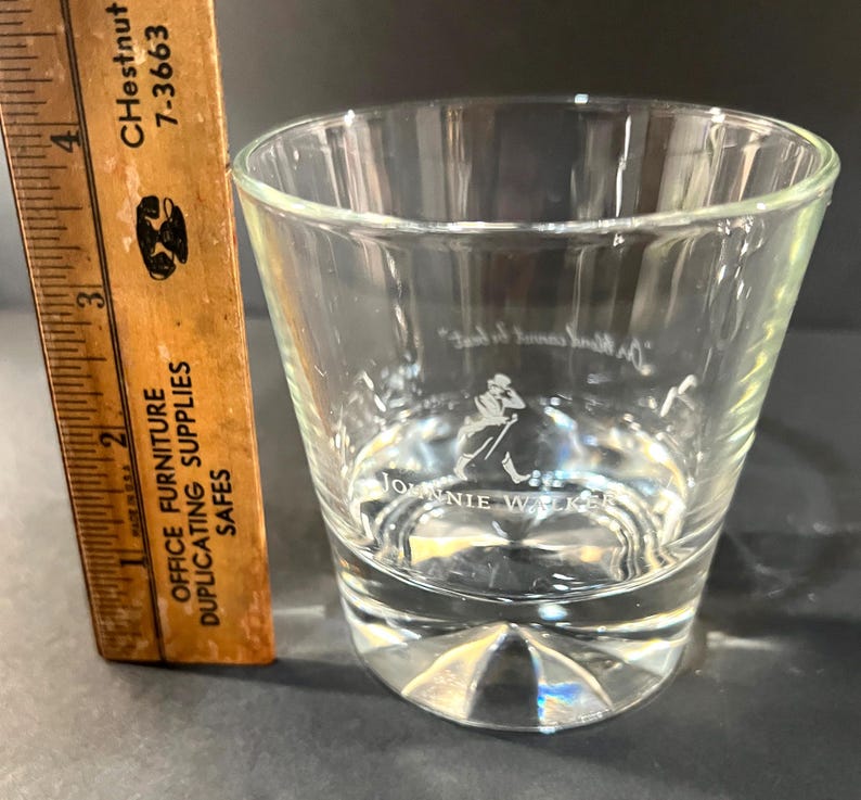 May include: A clear, lowball glass with the Johnnie Walker logo etched on the front. The glass has a thick base and a slightly tapered shape. A wooden ruler with inch markings is next to the glass.
