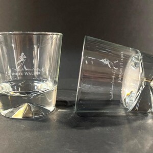 May include: Two clear, lowball whiskey glasses with the "Johnnie Walker" logo etched on the front. One glass is upright, the other is tilted on its side. The glasses have a thick, geometric base.