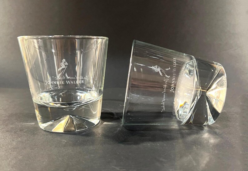 May include: Two clear, lowball whiskey glasses with the "Johnnie Walker" logo etched on the front. One glass is upright, the other is tilted on its side. The glasses have a thick, geometric base.
