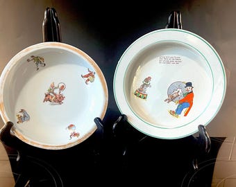Op de afbeelding: Twee antieke witte keramische borden met kleurrijke illustraties van circusartiesten. Eén bord heeft een bruine rand, de andere een groene rand en tekst. Beide borden worden op zwarte standaards gepresenteerd.
