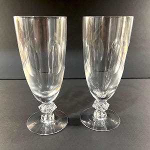 May include: Two clear, stemmed champagne flutes. The glasses have a tall, slender bowl with etched leaf-like designs. The base of each glass features a faceted, diamond-shaped detail above a circular foot. The flutes are displayed on a dark surface.