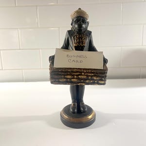 May include: A black and gold monkey figurine wearing a hat and jacket, standing on a circular base. The monkey holds a rectangular basket in front of its body, which contains a white card with the handwritten text BUSINESS CARD.