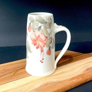 May include: A white ceramic beer stein with a large handle, decorated with a floral design in shades of pink, green, and brown. The stein is displayed on a wooden cutting board.