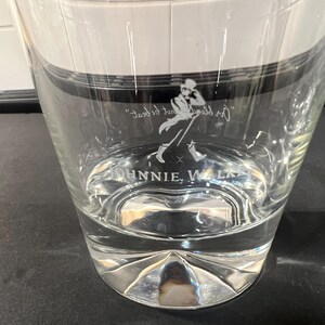 May include: Clear glass tumbler with a silver base. The glass features the Johnnie Walker logo and text. The tumbler is partially filled with a clear liquid. The glass has a unique, faceted base.