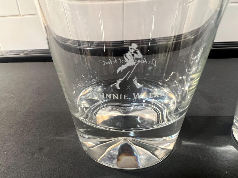 May include: Clear glass tumbler with a silver base. The glass features the Johnnie Walker logo and text. The tumbler is partially filled with a clear liquid. The glass has a unique, faceted base.