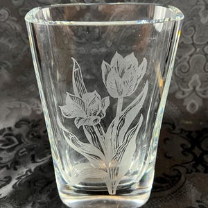 Op de afbeelding: Een heldere glazen vaas met een ge&euml;tst ontwerp van twee tulpen met gedetailleerde bloemblaadjes en bladeren. De vaas heeft een taps toelopende, cilindrische vorm met een dikke, massieve voet en gladde, transparante wanden met een gravure.