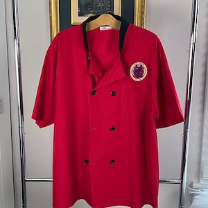 May include: Red short-sleeved chef jacket with a black collar and a black button closure. The jacket has a pocket on the left side and an embroidered logo on the right side that says "The Force" with a graphic of a bird.