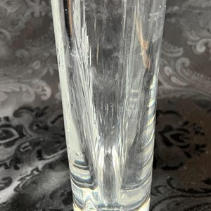 Op de afbeelding: Een heldere glazen vaas met een dikke, zware voet en een cilindervormig lichaam. Het glas is voorzien van verticale, getextureerde inkepingen die een gefacetteerd uiterlijk cre&euml;ren en het licht door de transparante structuur weerkaatsen.