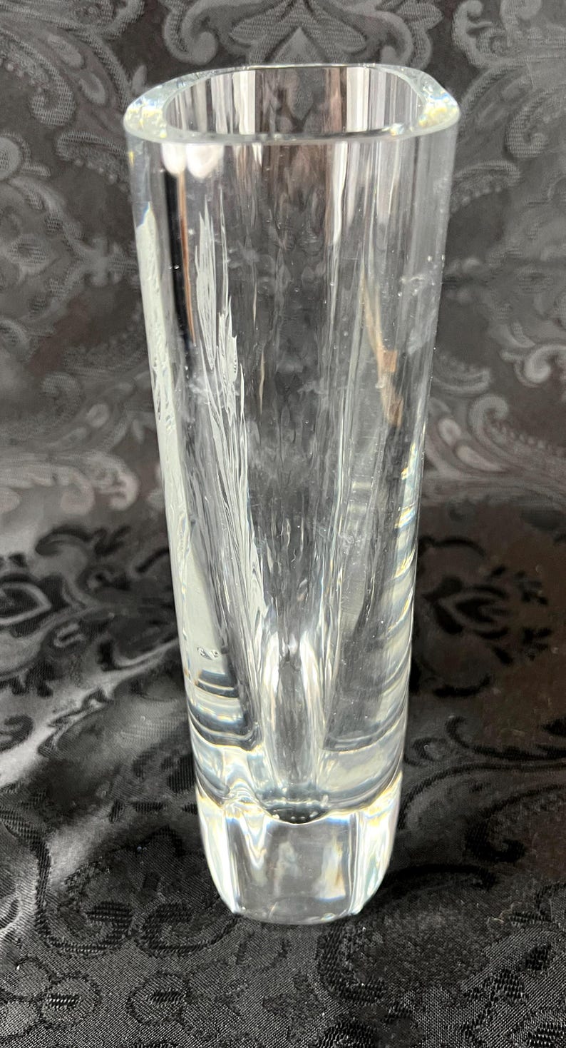 Op de afbeelding: Een heldere glazen vaas met een dikke, zware voet en een cilindervormig lichaam. Het glas is voorzien van verticale, getextureerde inkepingen die een gefacetteerd uiterlijk cre&euml;ren en het licht door de transparante structuur weerkaatsen.