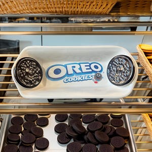 May include: A display of Oreo cookies in a bakery setting. A white tray with the Oreo logo and two large Oreo cookies is the focal point. Below, a tray of cookies and baskets of cookies are visible.