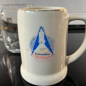 May include: A white ceramic mug with a gold rim and a blue and white image of the Space Shuttle Columbia. The image includes the text "Columbia Young-Crippen".