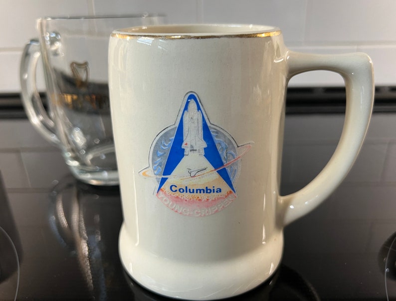 May include: A white ceramic mug with a gold rim and a blue and white image of the Space Shuttle Columbia. The image includes the text "Columbia Young-Crippen".