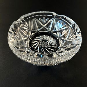 May include: A clear, cut-crystal ashtray with a starburst pattern in the center and geometric designs on the sides. The ashtray has a round shape with a small notch for resting cigarettes. The ashtray is made of glass.