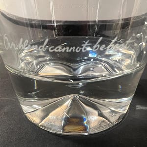 May include: A clear glass with a decorative, faceted base and the etched phrase "Our blind cannot be beat." The glass is partially filled with a clear liquid, likely water. The glass sits on a dark surface.