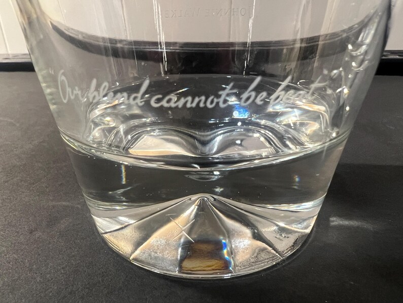 May include: A clear glass with a decorative, faceted base and the etched phrase "Our blind cannot be beat." The glass is partially filled with a clear liquid, likely water. The glass sits on a dark surface.